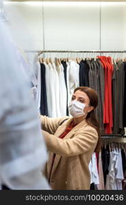 Beautiful girl wearing protective medical mask and fashionable clothes looks at clothes at store. New normal lifestyle concept.. Portrait of beautiful girl wearing protective medical mask