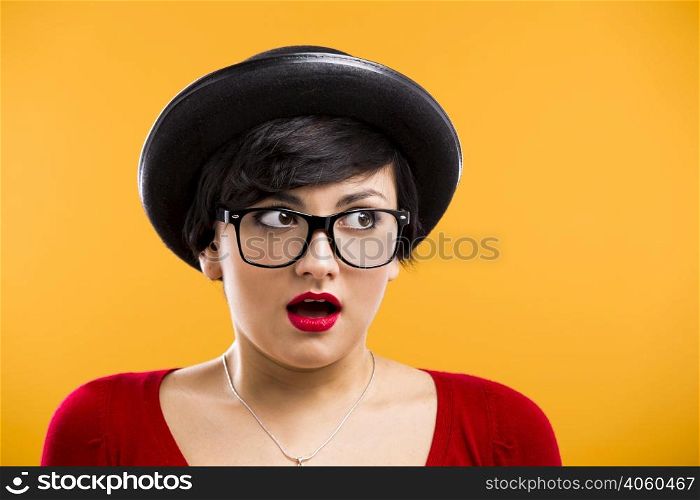 Beautiful girl with a astonished expression, wearing a hat and nerd glasses over a yellow background