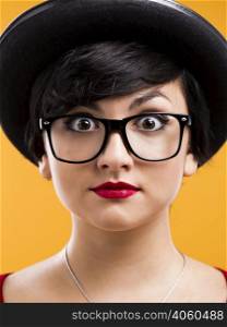 Beautiful girl with a astonished expression, wearing a hat and nerd glasses over a yellow background