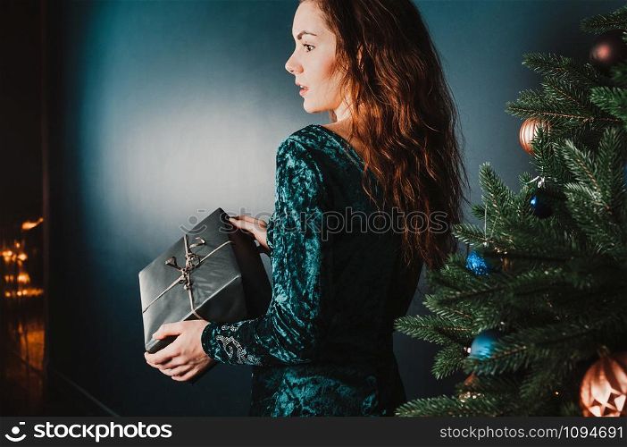 Beautiful girl with a gift box near Christmas tree, surprised look. New Year and Christmas concept. Copy space for your text and design. Luxury green, blue, golden colors