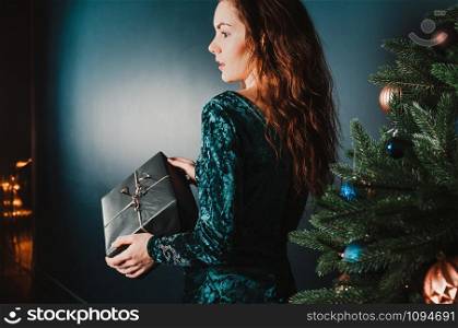 Beautiful girl with a gift box near Christmas tree, surprised look. New Year and Christmas concept. Copy space for your text and design. Luxury green, blue, golden colors