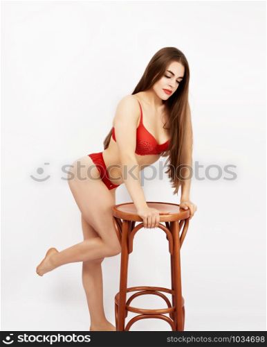 beautiful girl with a sports figure dressed in red lace underwear stands in a sideways position and rests on a brown wooden chair, white background