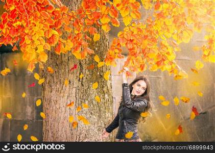 Beautiful girl with blue eyes, dressed in a black jacket, reaches the colorful branches, watching the falling leaves, enjoying the autumn atmosphere.