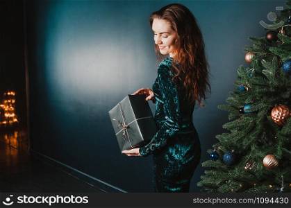 Beautiful girl with gift box near Christmas tree, smiling. New Year and Christmas concept. Copy space for your text and design.