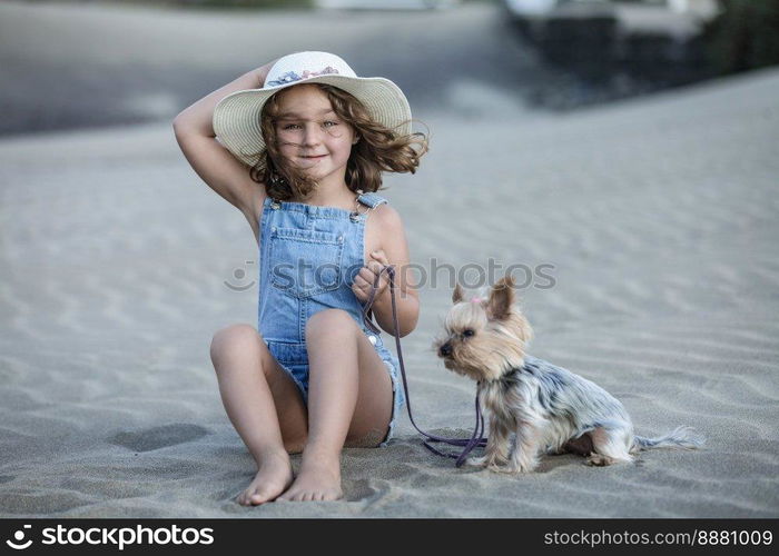 Beautiful girl with hat looking straight ahead sitting on the beach sand holding the leash of her pet yorkshire terrier.