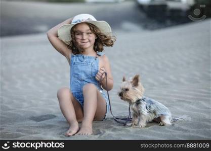 Beautiful girl with hat looking straight ahead sitting on the beach sand holding the leash of her pet yorkshire terrier.