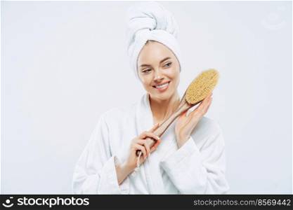 Beautiful glad woman with healthy skin and toothy smile holds wooden massage brush for body, wears bath robe and wrapped soft towel on head, stands against white background. Beauty, care concept