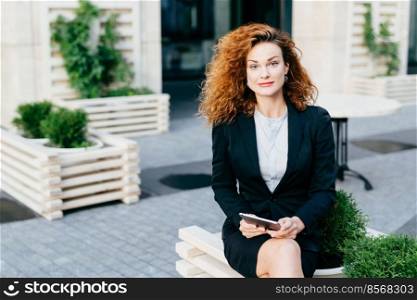 Beautiful gorgeous woman with wavy bushy hair, wearing formal suit, sitting crossed legs at outdoor cafe, holding tablet computer while waiting for someone, looking at camera with confident look