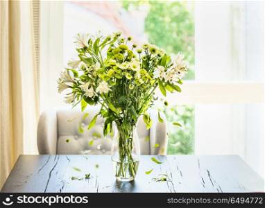 Beautiful green flowers bunch with falling petals in glass vase on table in sunny living room at window. Home interior and decor ideas