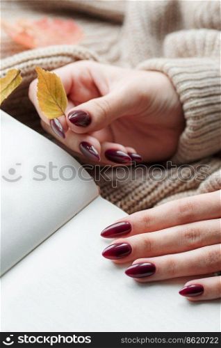 Beautiful hands of a young woman with dark red manicure on nails. Autumn concept. Hands of a girl in a warm brown sweater leafing through a book