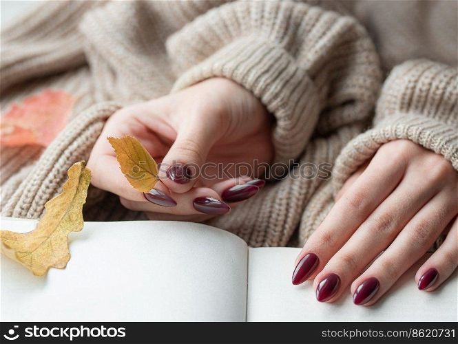 Beautiful hands of a young woman with dark red manicure on nails. Autumn concept. Hands of a girl in a warm brown sweater leafing through a book