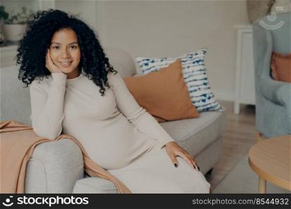 Beautiful happy african expectant mother sitting in relaxed pose while being pregnant, resting head on hand leaning on side of cozy sofa in living room, smiling and enjoying her leisure time at home. Beautiful african expectant mother sitting in relaxed pose in living room