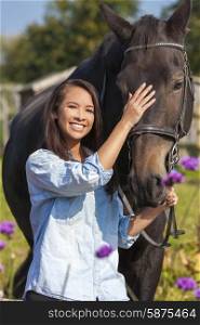 Beautiful happy Asian Eurasian young woman or girl wearing denim shirt, smiling and leading her horse in sunshine