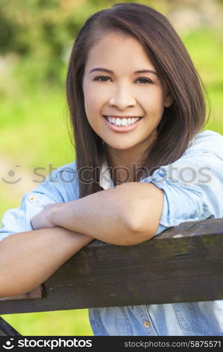 Beautiful happy Asian Eurasian young woman or girl wearing denim shirt, smiling and leaning on fence in sunshine