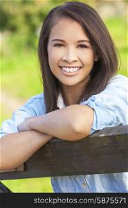 Beautiful happy Asian Eurasian young woman or girl wearing denim shirt, smiling and leaning on fence in sunshine