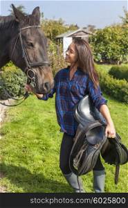 Beautiful happy Asian Eurasian young woman or girl wearing plaid checked shirt, csrrying a saddle to her horse in sunshine