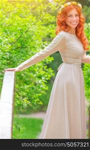 Beautiful happy bride wearing long beige dress and flower wreath in red hair, standing on little bridge in fresh summer park, happiness and love concept