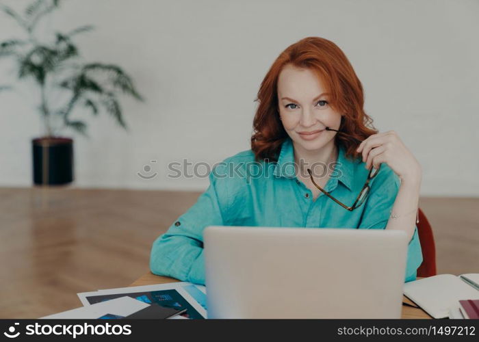 Beautiful happy busy female freelance worker with ginger hair, works online, uses laptop computer, prepares financial report at home, uses videoconferencing app, talks distantly with colleagues