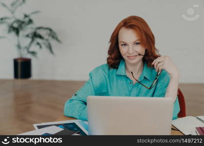 Beautiful happy busy female freelance worker with ginger hair, works online, uses laptop computer, prepares financial report at home, uses videoconferencing app, talks distantly with colleagues