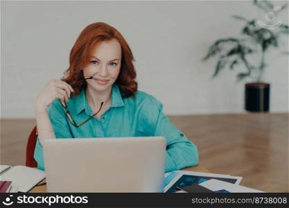 Beautiful happy busy female freelance worker with ginger hair, works online, uses laptop computer, prepares financial report at home, uses videoconferencing app, talks distantly with colleagues