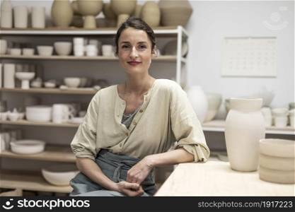 Beautiful happy craft woman wearing apron looking at camera and smiling while sitting in her art studio or craft pottery shop. Portrait of beautiful happy craft woman wearing apron and smiling while sitting in her art studio or craft pottery shop