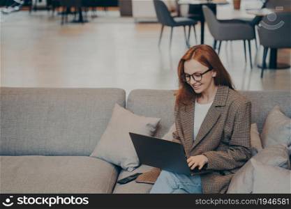 Beautiful happy female employee in casual clothes sitting on comfortable couch in office break room, using laptop to browse web, smiling and feeling satisfied, furniture in blurred background. Happy female employee browsing web while sitting on couch in break room in modern office