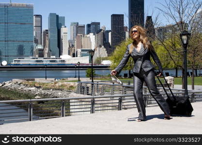 Beautiful happy laughing Caucasian Latina entrepreneur business woman in grey suit running with a carry-on luggage and purse through park in New York City.