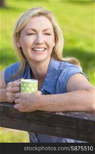 Beautiful happy middle aged woman in her thirties or forties smiling and leaning on fence in sunshine drinking tea or coffee