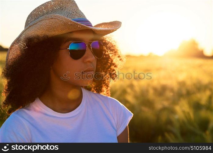 Beautiful happy mixed race African American female girl teenager young woman wearing relective aviator sunglasses and cowboy hat in a cornfield at golden sunset or sunrise