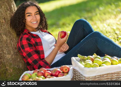 Beautiful happy mixed race African American girl teenager female young woman in an orchard eating an organic red apple, smiling with perfect teeth leaning against a tree with baskets of apples she has been picking