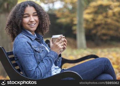 Beautiful happy mixed race African American girl teenager female young woman smiling and drinking takeaway coffee outside sitting on a park bench in autumn or fall