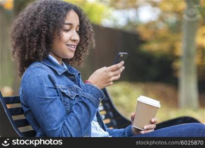 Beautiful happy mixed race African American girl teenager female young woman smiling drinking takeaway coffee and cell phone texting sitting on a park bench in autumn or fall