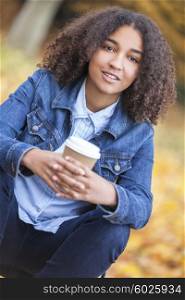 Beautiful happy mixed race African American girl teenager female young woman smiling and drinking takeaway coffee outside sitting on a park bench in autumn or fall