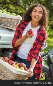 Beautiful happy mixed race African American girl teenager female young woman smiling with perfect teeth in an orchard resting on a tractor eating an apple with baskets of organic apples she has been picking