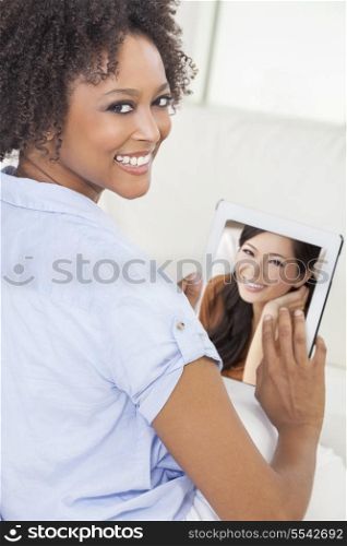 Beautiful happy mixed race African American woman or girl at home sitting on her sofa &amp; smiling using a tablet computer for a video call to her Asian Chinese female friend