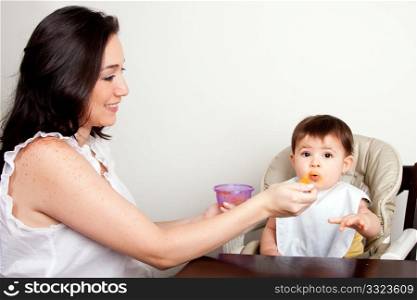 Beautiful happy mother or nanny feeds funny baby boy girl orange puree with spoon, infant eats messy, while sitting at table.