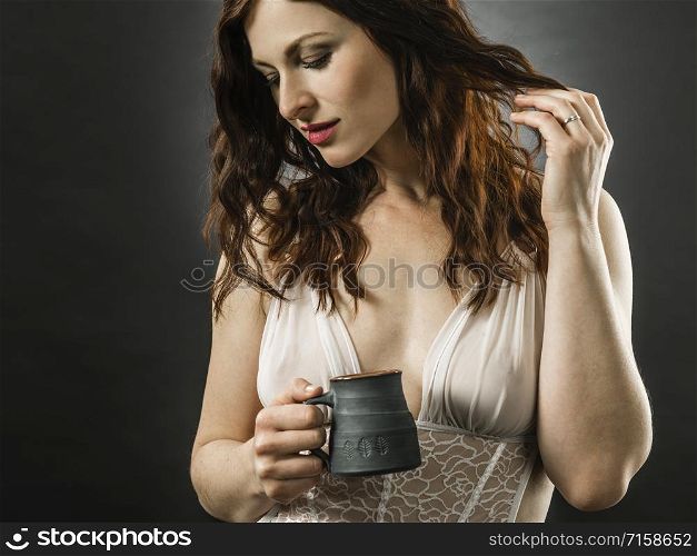 Beautiful happy redhead woman wearing lingerie and drinking coffee over dark background.
