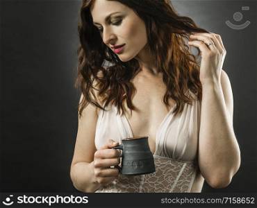 Beautiful happy redhead woman wearing lingerie and drinking coffee over dark background.