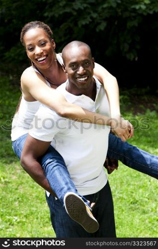 Beautiful happy smiling laughing African American couple piggyback playing in the park, woman hugging man, wearing white shirts and blue jeans.