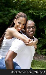 Beautiful happy smiling laughing young African American couple piggyback playing in the park, woman hugging man, wearing white shirts and blue jeans.