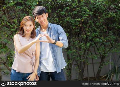 Beautiful happy young couple fun making gesture heart shape with hand outdoor together, man and woman with relation feeling love with symbol and sign in the garden, lover and romantic concept.