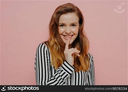 Beautiful happy young female in stripy black and white blouse touching lips with pointer finger while posing in studio with light pink background. Pretty smiling young girl looking at camera touching lips while standing against pink wall