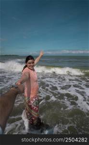 Beautiful happy young Hispanic woman holding her partner&rsquo;s hand at the beach, wearing a pink dress with the sea in the background during a sunny morning. Beautiful happy young Hispanic woman holding her partner&rsquo;s hand at the beach, wearing a pink dress during a sunny morning