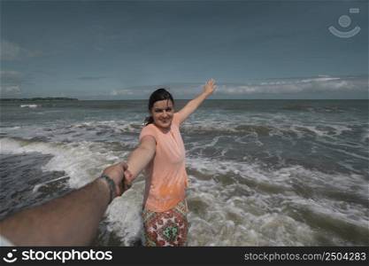 Beautiful happy young Hispanic woman holding her partner&rsquo;s hand at the beach, wearing a pink dress with the sea in the background during a sunny morning. Beautiful happy young Hispanic woman holding her partner&rsquo;s hand at the beach, wearing a pink dress during a sunny morning