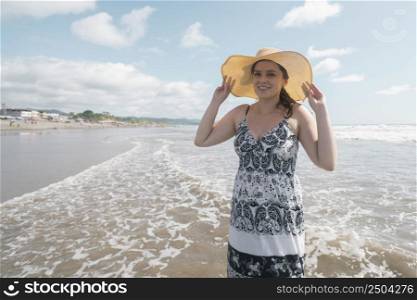 Beautiful happy young Hispanic woman walking alone on the beach holding her hat in her hands and wearing a black and white dress during a sunny morning. Beautiful happy young Hispanic woman walking alone on the beach wearing a hat and black and white dress during a sunny morning