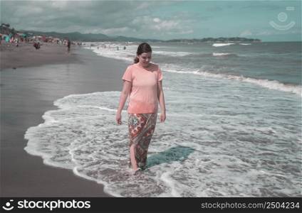 Beautiful happy young Hispanic woman walking alone on the beach stepping into the waves in a pink dress during a sunny morning. Beautiful young Hispanic woman walking alone on the beach in a pink dress during a sunny morning