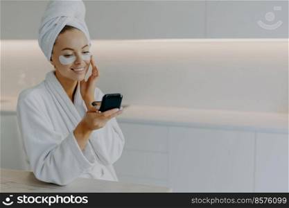 Beautiful happy young lady applies collagen patches under eyes looks at herself in mirror does rejuvenation procedure wears bath towel and dressing gown after taking shower has skin care routine
