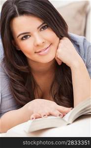 Beautiful happy young Latina Hispanic woman laying down reading a paperback book at home on her sofa