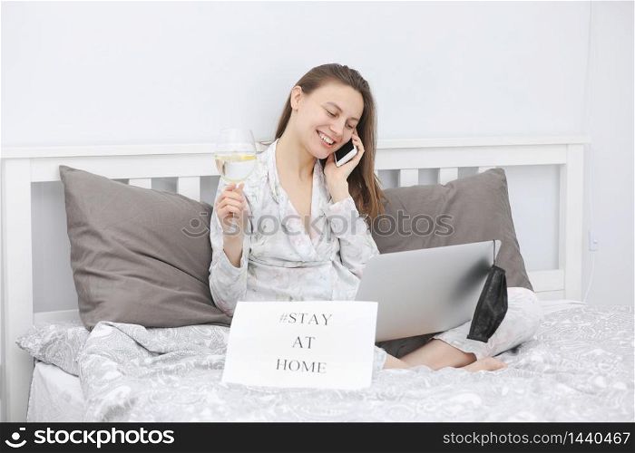 Beautiful happy young woman with a glass of water working on laptop computer in the bedroom. Stay at home. Freelancer. Writing, typing. Girl checking social apps. Communication and technology concept. Beautiful happy young woman with a glass of water working on laptop computer in the bedroom. Stay at home. Freelancer. Writing, typing. Girl checking social apps. Communication and technology concept.
