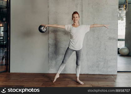 Beautiful healthy ginger woman performing barre exercises with small fitball, standing in second position with hands up in fitness club or studio, dressed in sportswear. Sport and pilates concept. Slender beautiful red haired woman performing barre exercises in fitness studio